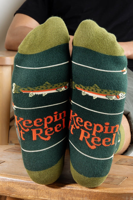 Green fishing socks with 'Keeping it Reel' text worn by a person sitting on a wooden stool.