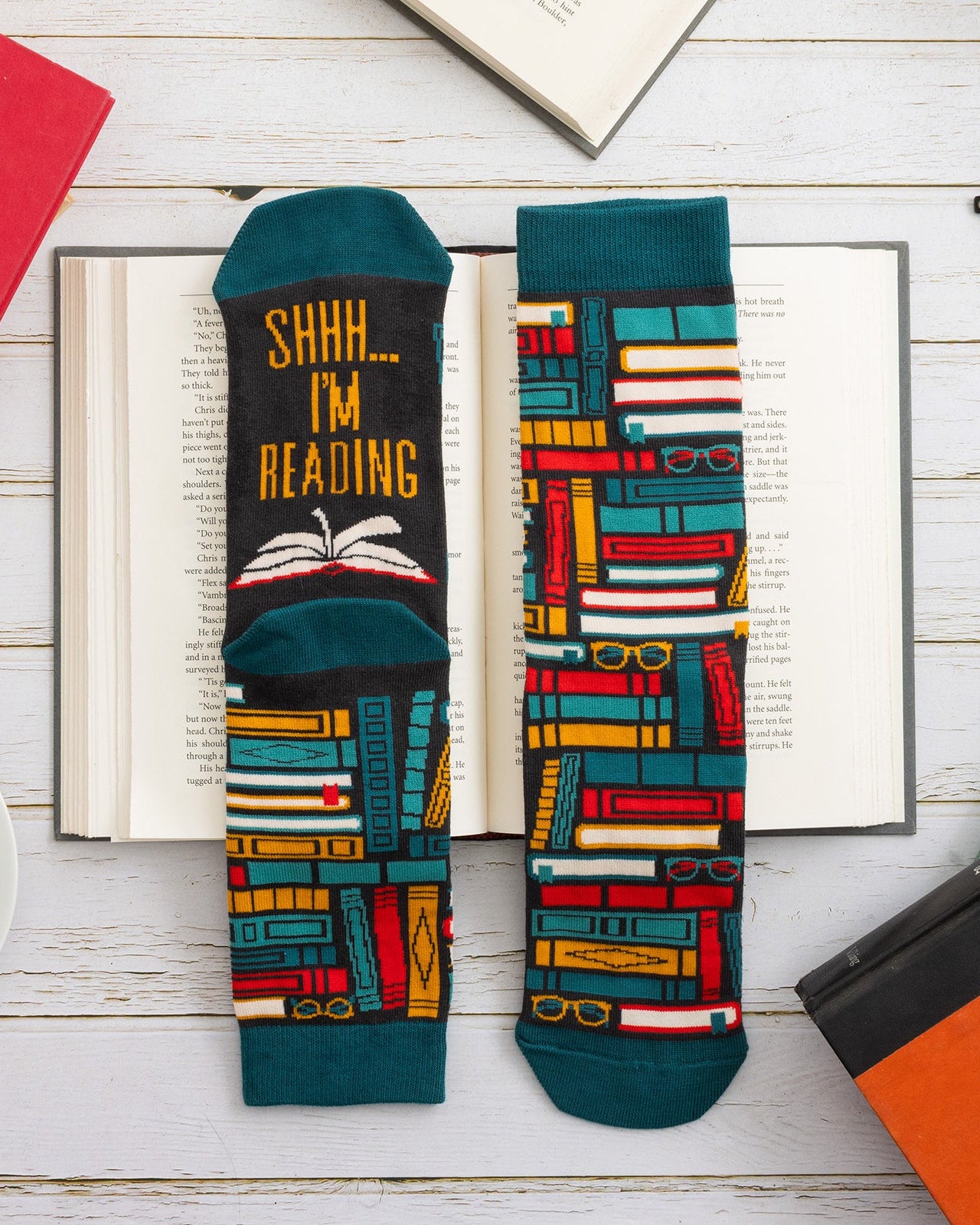 Two socks with book-themed designs on a wooden surface with books and a cup.
